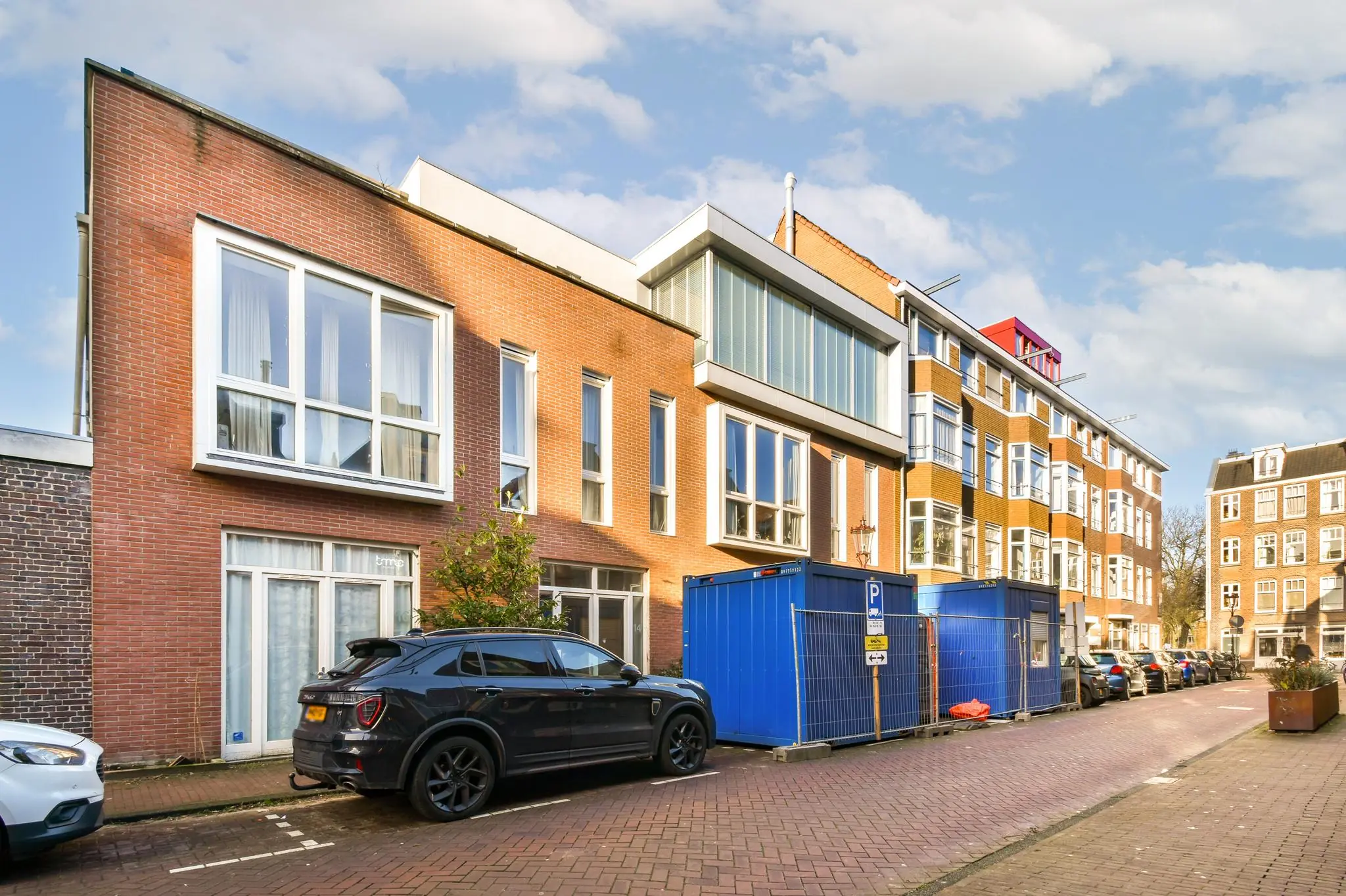 Straatbeeld van de Korte Schimmelstraat met moderne huizen, geparkeerde auto’s en blauwe bouwcontainers.